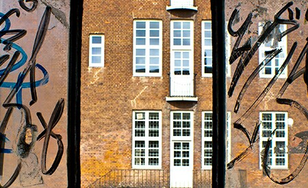 Ventana de 3 paneles que da a un edificio de ladrillo marrón. Los vidrios laterales tienen grafitis negros y el central es transparente y limpio.

