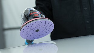 Mano con guante y lijadora orbital 3M con disco perforado sobre una superficie lisa.
